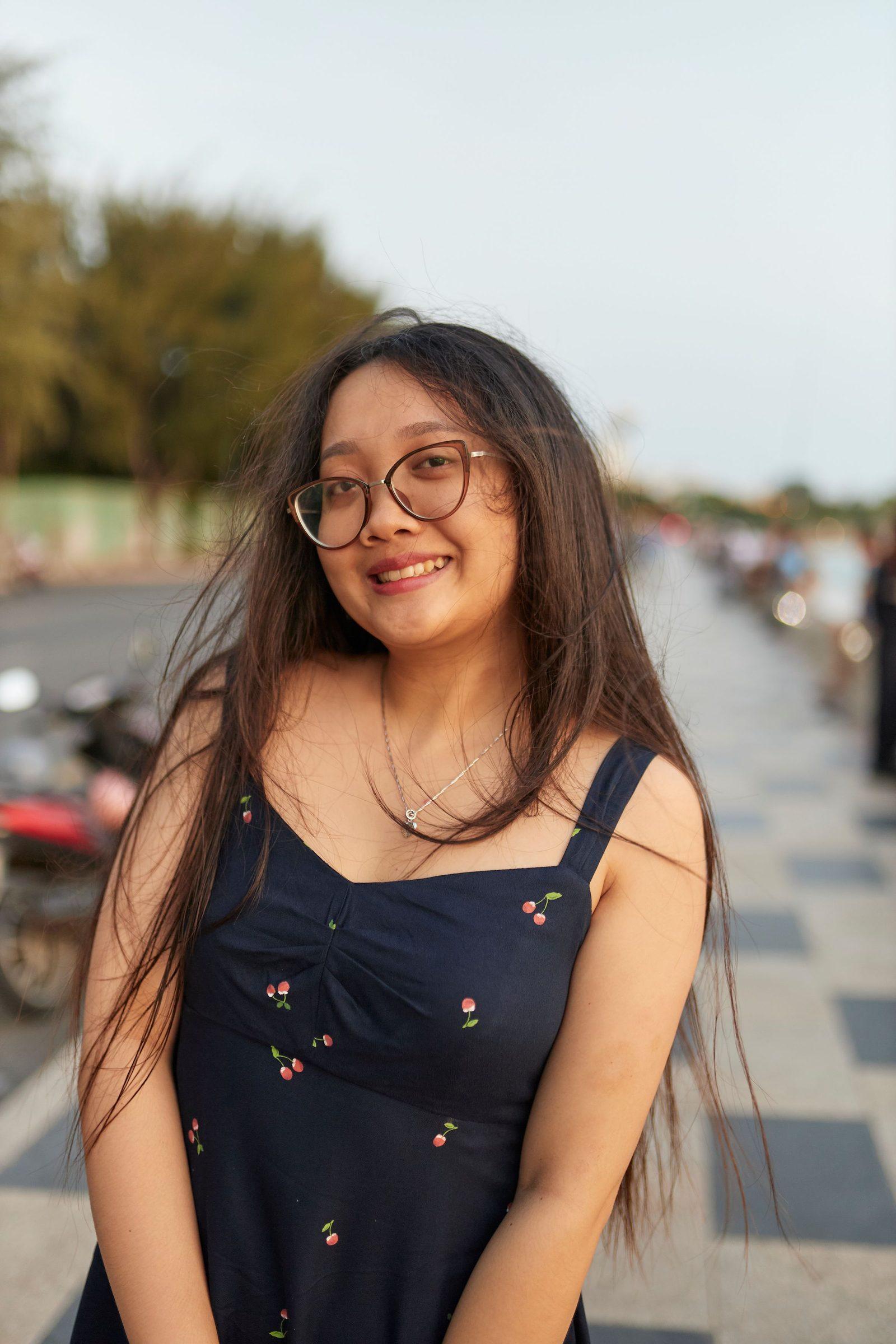Cô gái tóc dài đeo gọng kính cười trên con đường ven biển
